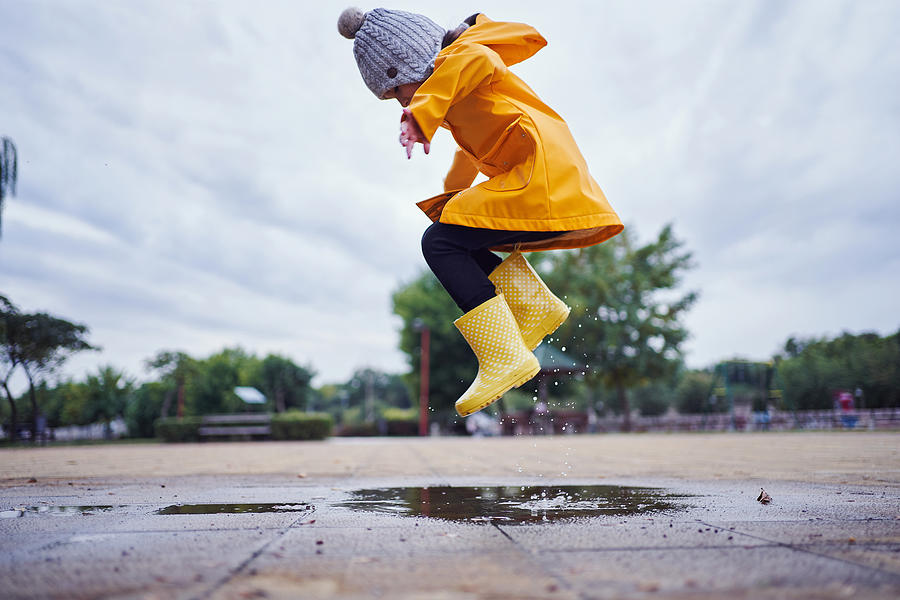 Mid-air shot of a child jumping in a puddle of water wearing yellow rubber boots and a raincoat in autumn Photograph by FluxFactory