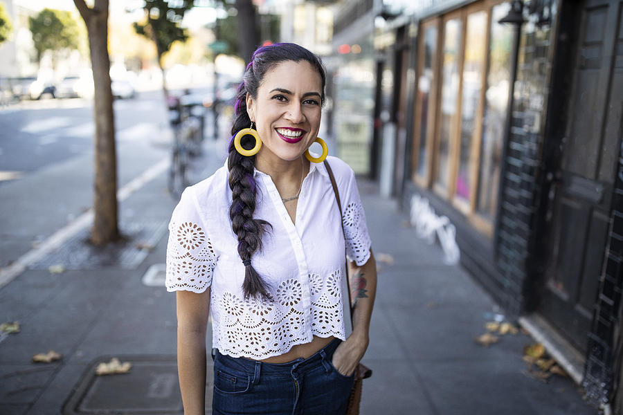 Millennial Latina Stands Alone on City Sidewalk, Smiling and Looking at Camera, Wearing White Lace Blouse and Bright Yellow Hoop Earrings Photograph by Justin Lewis