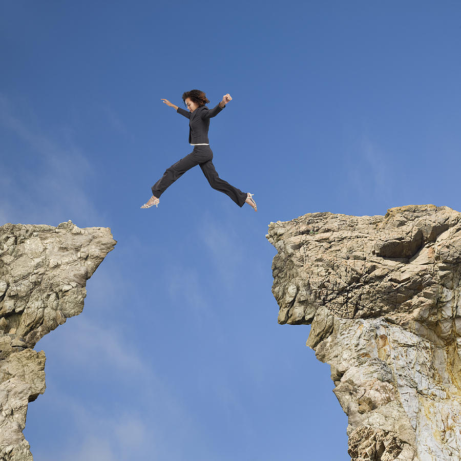 Mixed race businesswoman jumping over gap between cliffs Photograph by John M Lund Photography Inc