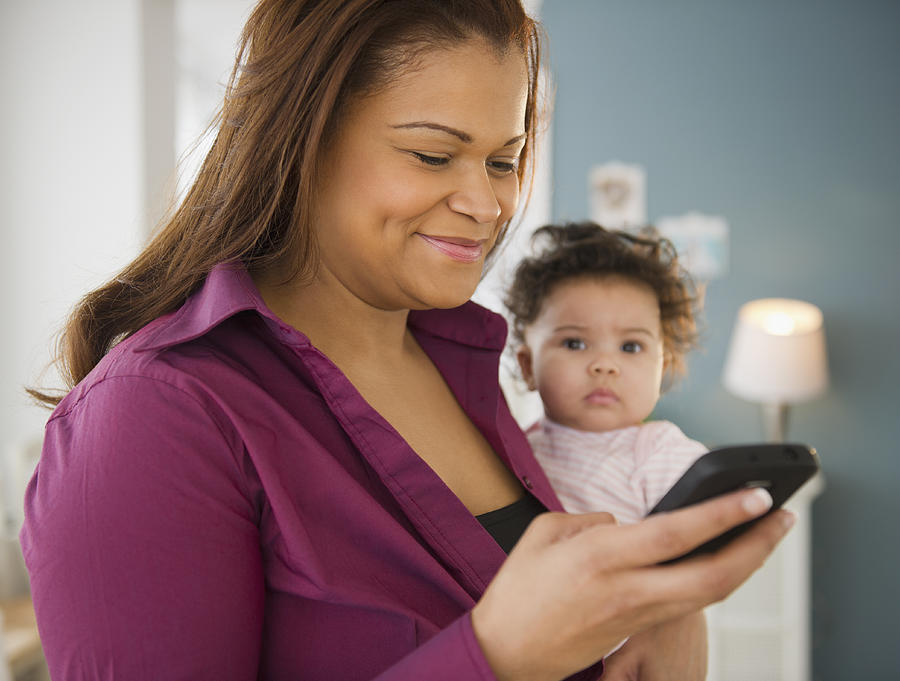 Mixed race mother holding baby and text messaging on cell phone Photograph by Blend Images - JGI/Jamie Grill