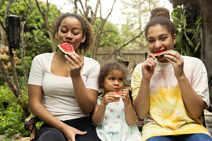 Mixed-race sisters eating watermelon in backyard. Photograph by Martinedoucet