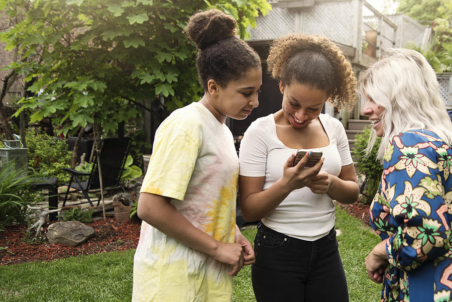 Mixed-race sisters looking at mobile phone with mother in backyard. Photograph by Martinedoucet