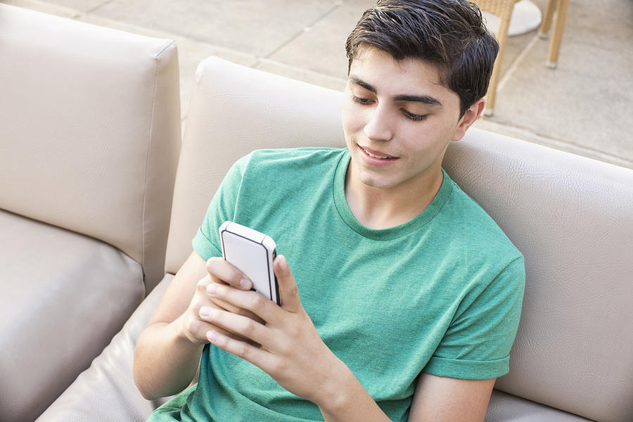 Mixed race teenage boy using cell phone on sofa Photograph by Kingfisher Productions