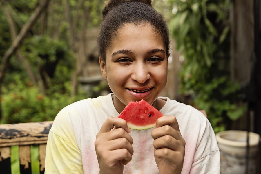 Mixed-race teenage girl with braces eating watermelon in backyard. Photograph by Martinedoucet