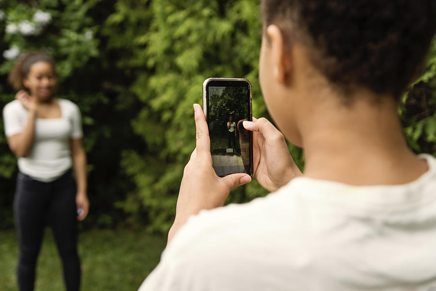 Mixed-race teenage sisters filming with mobile phone in backyard. Photograph by Martinedoucet
