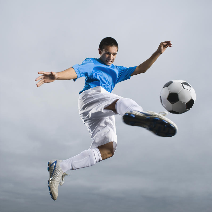 Mixed race teenager in mid-air kicking soccer ball Photograph by Blend Images - Erik Isakson
