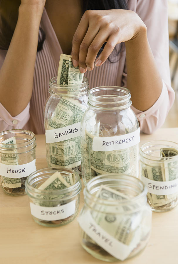 Mixed race woman putting money in savings jars Photograph by JGI/Jamie Grill