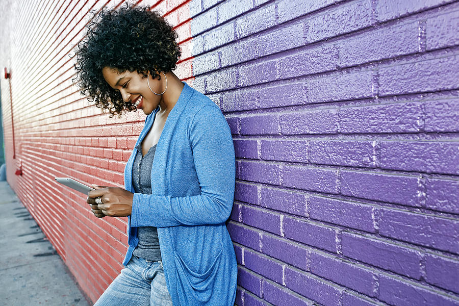Mixed race woman with cell phone standing by colorful wall Photograph by Peter Griffith