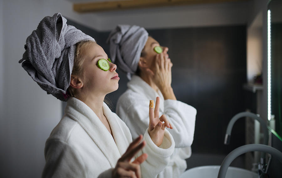 Mother and small daughter indoors in bathroom at home, putting cucumbers on eyes. Photograph by Halfpoint Images