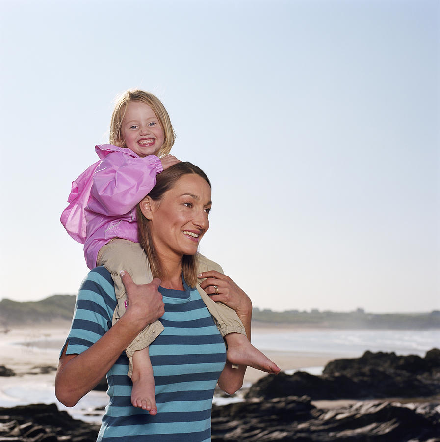 Mother carrying daughter (2-4) on shoulders by beach, smiling Photograph by Janie Airey