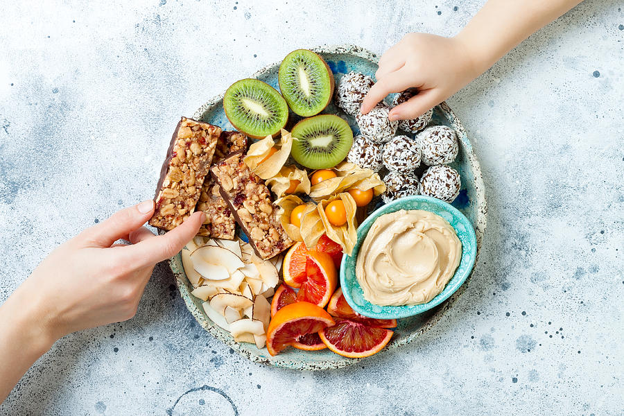 Mother giving healthy vegan dessert snacks to toddler child. Concept of healthy sweets for children. Protein granola bars, homemade raw energy balls, cashew butter, toasted coconut chips, fruits platter Photograph by Sveta_zarzamora