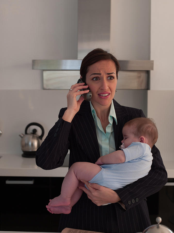 Mother holding baby daughter while talking on phone in kitchen Photograph by Chris Ryan