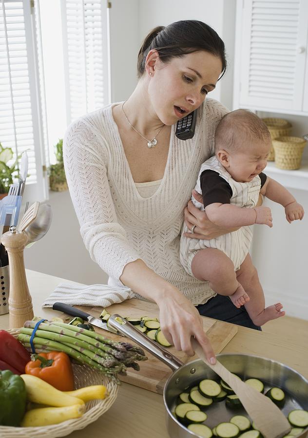 Mother holding baby, talking on telephone and cooking Photograph by Tetra Images