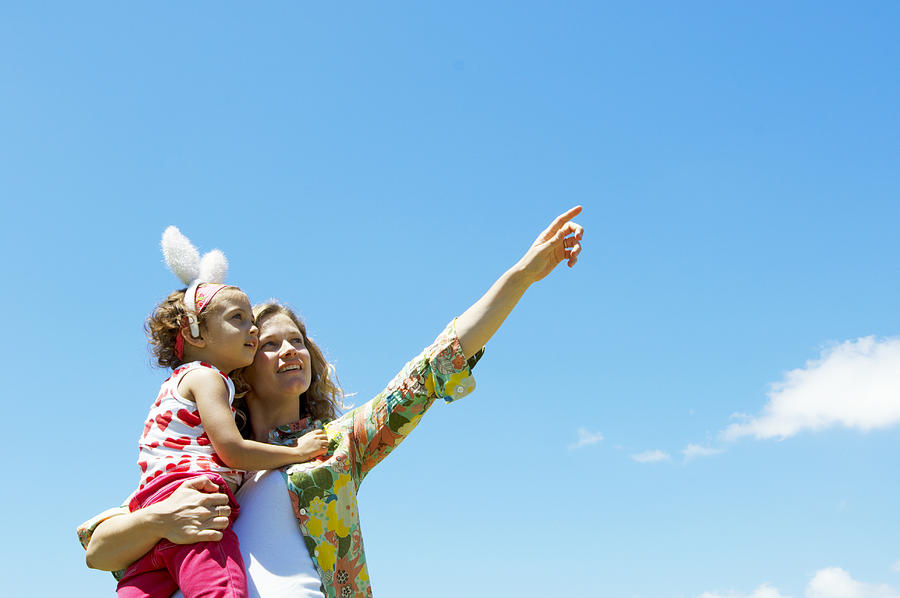 Mother holding daughter (3-5) wearing bunny ears, pointing upwards Photograph by Kane Skennar