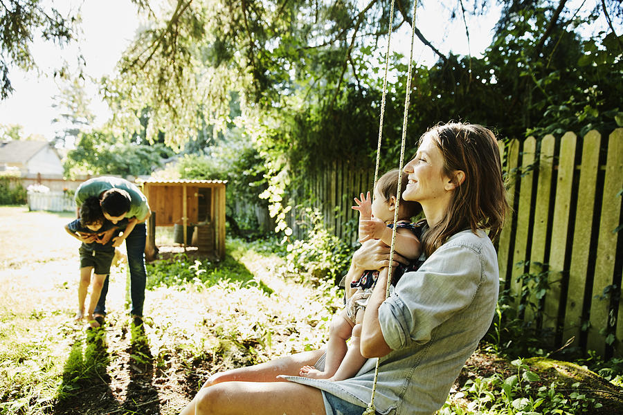 Mother holding infant daughter on swing while father plays with son in backyard Photograph by Thomas Barwick