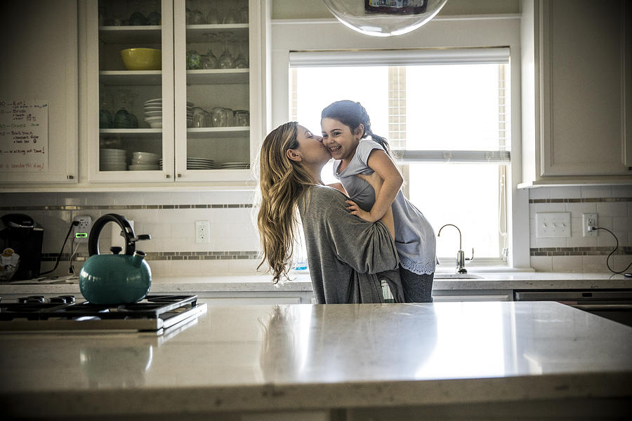 Mother kissing daughter (7yrs) in kitchen Photograph by MoMo Productions