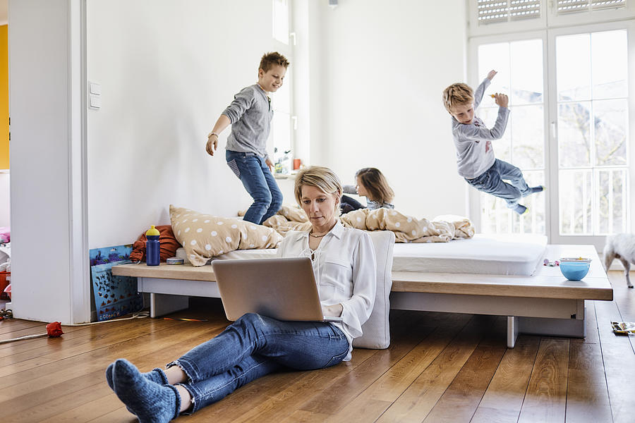 Mother using laptop at home with children romping around in background Photograph by Oliver Rossi