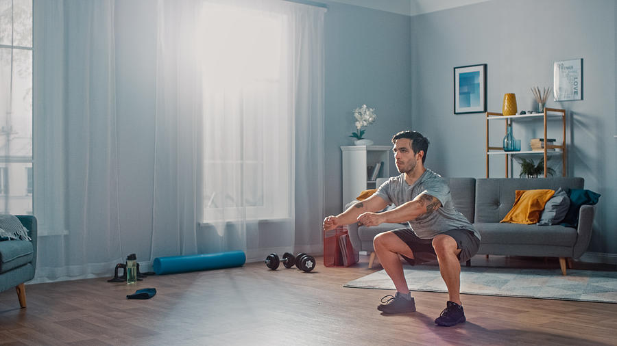Muscular Athletic Fit Man in T-shirt and Shorts is Doing Squat Exercises at Home in His Spacious and Bright Living Room with Minimalistic Interior. Photograph by Gorodenkoff