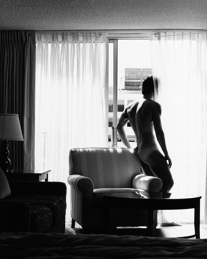 Naked man looking out window, rearview (B&W) Photograph by Dana Menussi