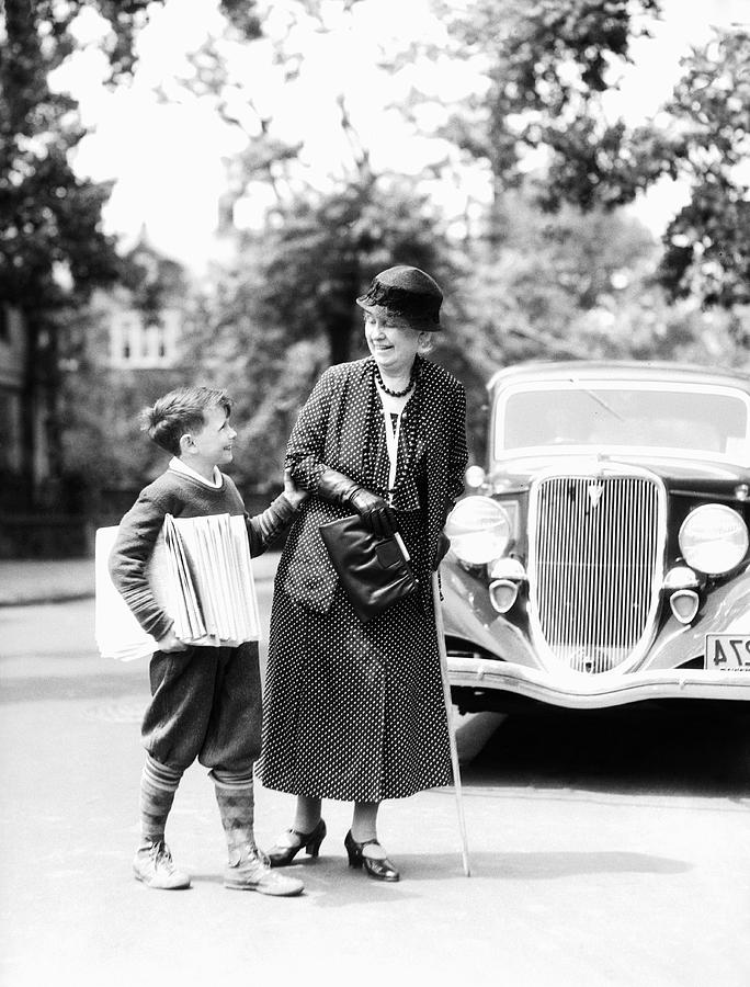 Newsboy with stack of papers under arms, helping elderly woman with cane cross street. Photograph by H. Armstrong Roberts