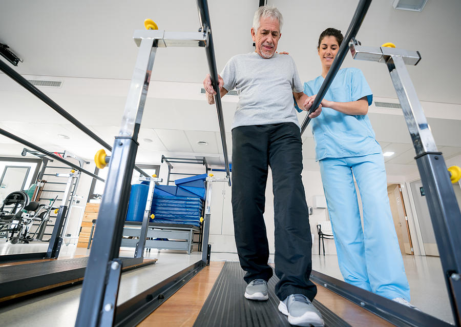 Occupational therapist helping senior patient on his recovery using parallel bars to walk Photograph by Andresr