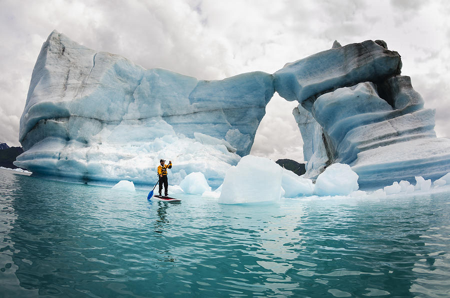 One man on stand up paddle board (SUP) paddles past hole melted in iceberg on Bear Lake in Kenai Fjords National Park, Alaska. Photograph by James + Courtney Forte