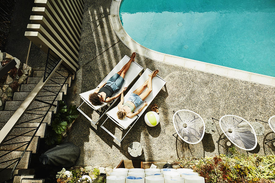 Overhead view of lesbian couple holding hands while relaxing in lounge chairs by hotel pool Photograph by Thomas Barwick