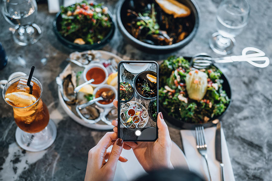 Overhead view of young woman taking photos of scrumptious and delicious meal on dining table with smartphone before eating it in restaurant. Eating out lifestyle. Camera eats first culture Photograph by D3sign