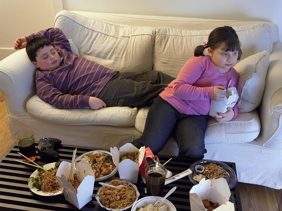 Overweight Brother and Sister on a Sofa Eating Takeaway Food and Watching the TV Photograph by Digital Vision.