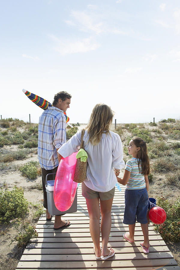 Parents and daughter (6-8) on beach path carrying picnic box and toys Photograph by Getty Images