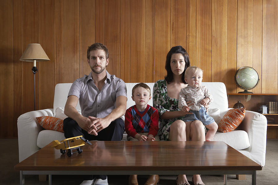 Parents with son (3-5), and baby girl (6-9 months) sitting on couch in living room Photograph by Noel Hendrickson