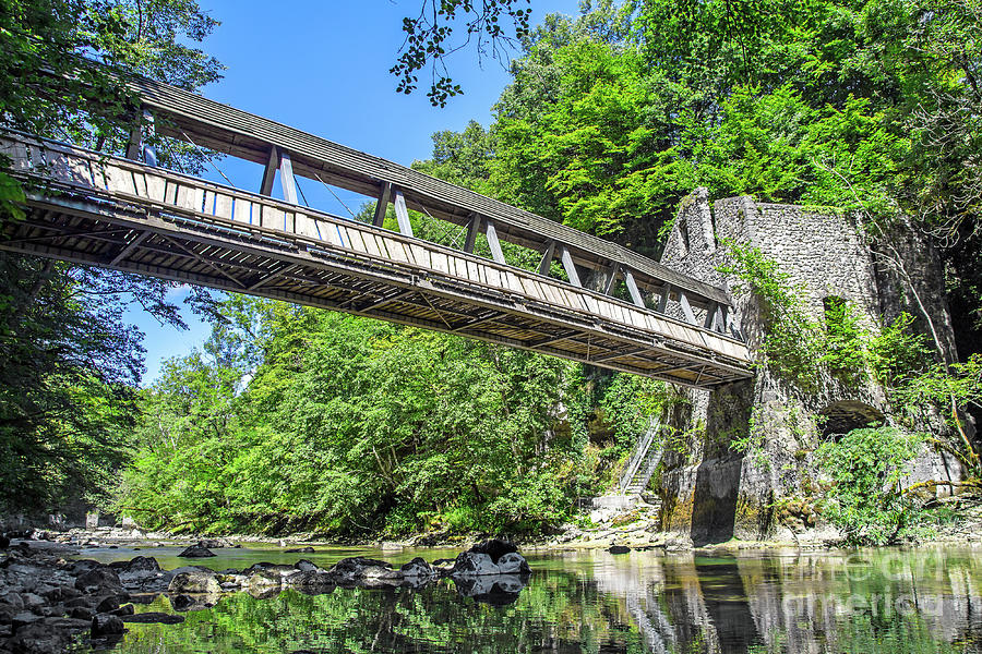 Pedestrian bridge crossing wild river with old mill building Photograph by Gregory DUBUS