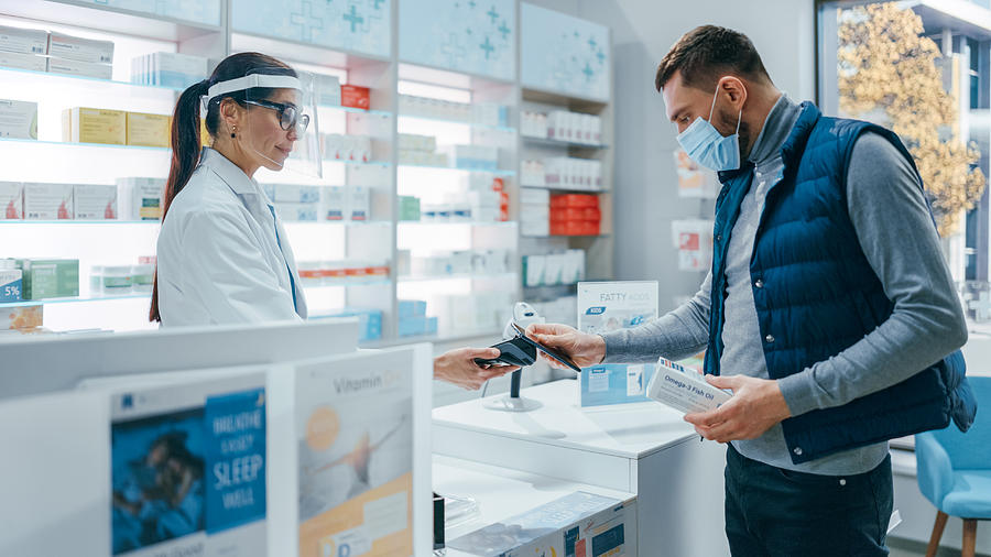 Pharmacy Drugstore Checkout Counter: Female Pharmacist Wearing Face Shields Selling Medicine Package, Customer with Face Mask Using NFC Smartphone with Contactless Payment Terminal and Credit Card Photograph by Gorodenkoff