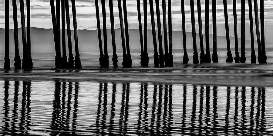 Black And White Photograph - Pismo Beach Pier Piling Silhouettes and Coastal Mountain Panorama - Black and White  by Gregory Ballos