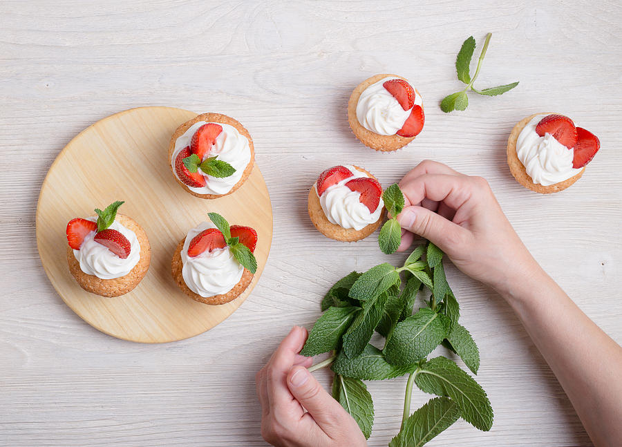 Placing mint leaf on the top of a buttercream cupcake with strawberries viewed from above Photograph by Istetiana