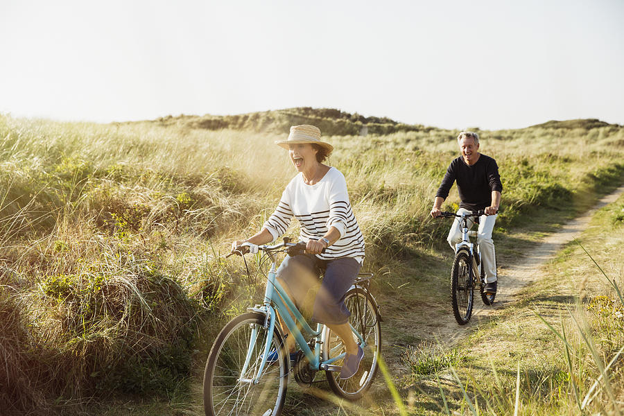 Playful mature couple riding bicycles on sunny beach grass path Photograph by Caia Image
