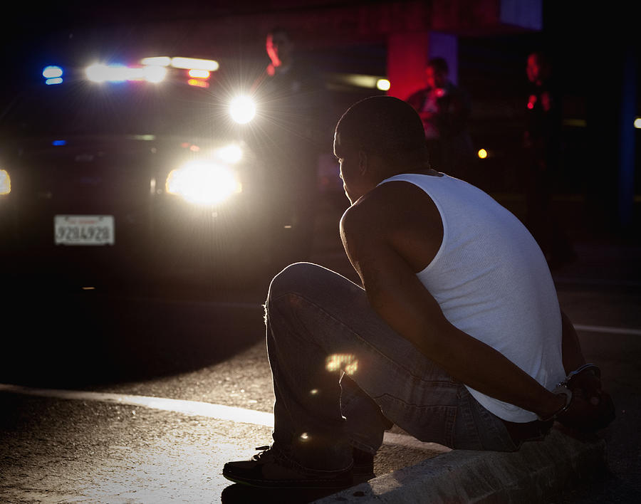 Police shining lights on handcuffed African man sitting on curb Photograph by Hill Street Studios