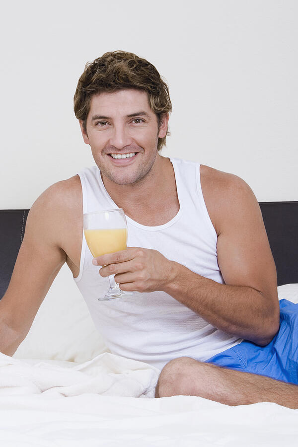 Portrait of a mid adult man sitting on the bed and holding a glass of orange juice Photograph by Glowimages
