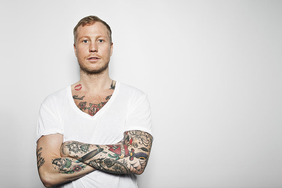 Portrait of a tattooed man with arms crossed standing against grey background Photograph by Maskot
