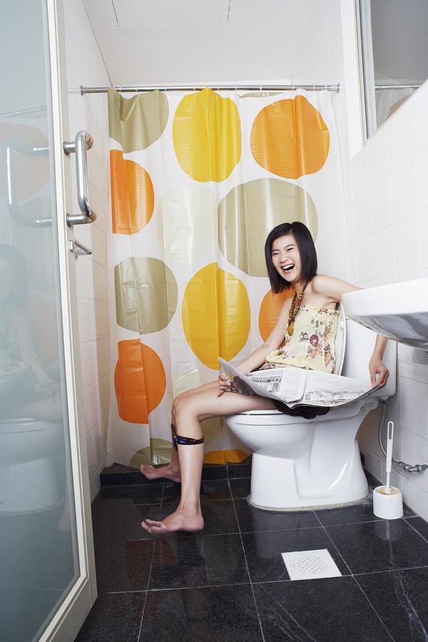 Portrait of a young woman sitting on a toilet bowl and holding a newspaper Photograph by Red Chopsticks