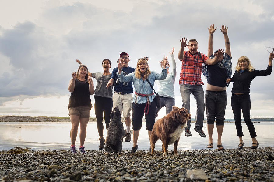Portrait of adult family with dogs jumping on shingle beach, Maine, USA Photograph by Heshphoto