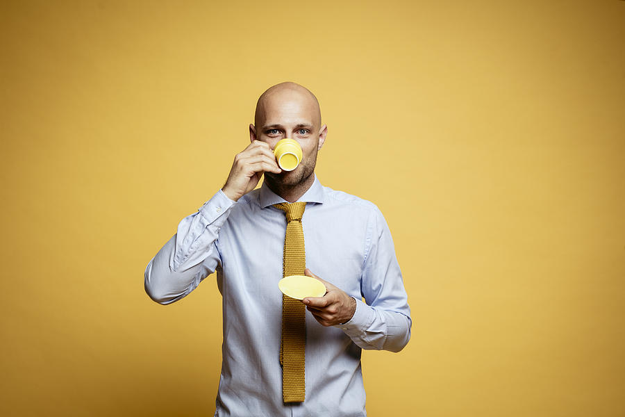 Portrait of bald businessman drinking cup of coffee against yellow background Photograph by Westend61