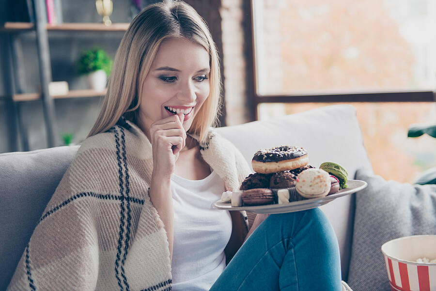 Portrait of beautiful emotional charming attractive sweet toothy woman sitting on sofa in living room, holding plate of donuts and macaroons, looking exciting satisfied Photograph by Deagreez