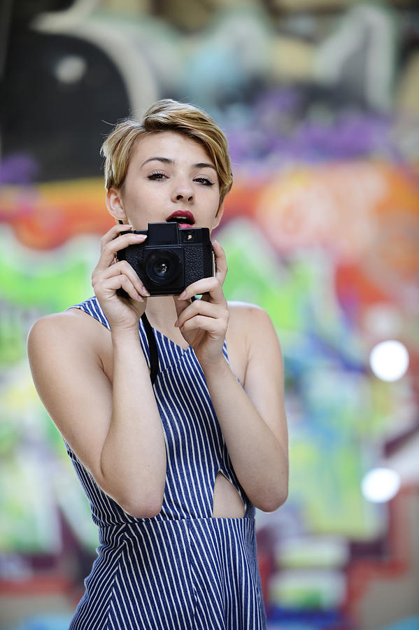 Portrait of cool teenage girl holding up camera in front of graffiti wall Photograph by Pete Saloutos