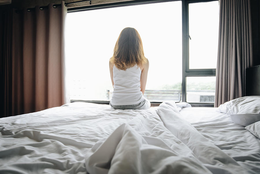 Portrait of depressed woman sitting alone on bed, looking to outside the window. Photograph by Boy_Anupong