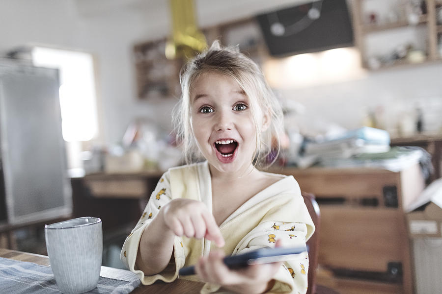 Portrait of excited little girl in the kitchen pointing at smartphone Photograph by Westend61