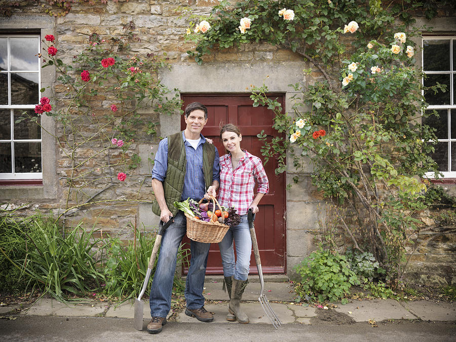 Portrait of farming couple with basket of organic vegetables and gardening tools standing in front of farmhouse doorway Photograph by Monty Rakusen