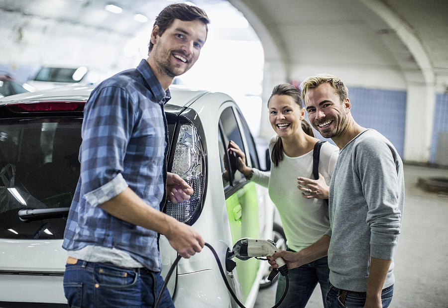 Portrait of happy friends with man charging electric car at gas station Photograph by Maskot