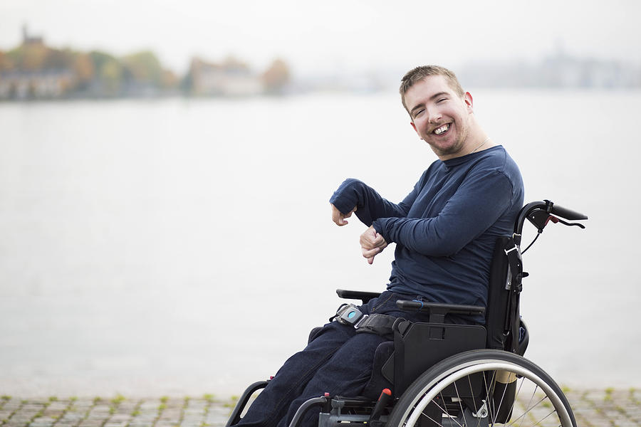 Portrait of happy man with cerebral palsy sitting on wheelchair by lake Photograph by Maskot