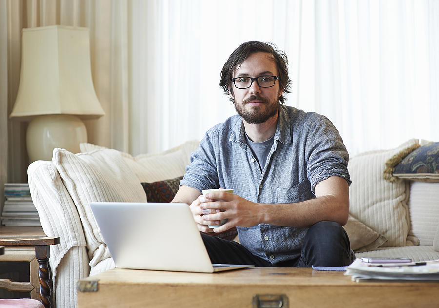 Portrait of man sitting on sofa with laptop and hot drink. Photograph by Dougal Waters